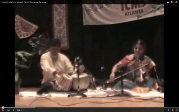 Two musicians sitting on the floor playing traditional Indian string instruments, with a banner and decorative textiles in the background.