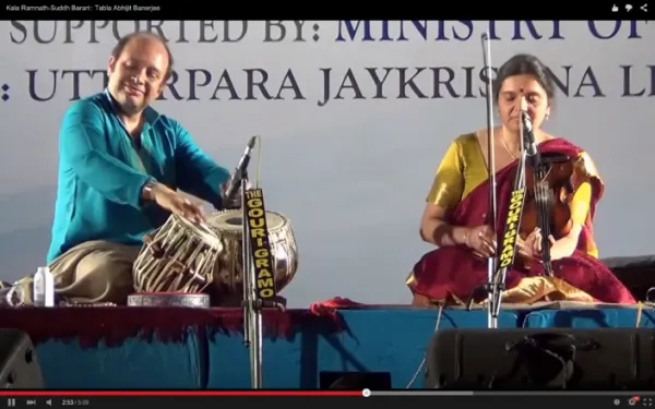 A man and a woman are performing traditional Indian music on stage. The man plays a tabla, and the woman plays a string instrument. They are sitting on a stage with a blue cloth covering the platform, and there is a large banner in the background with tex