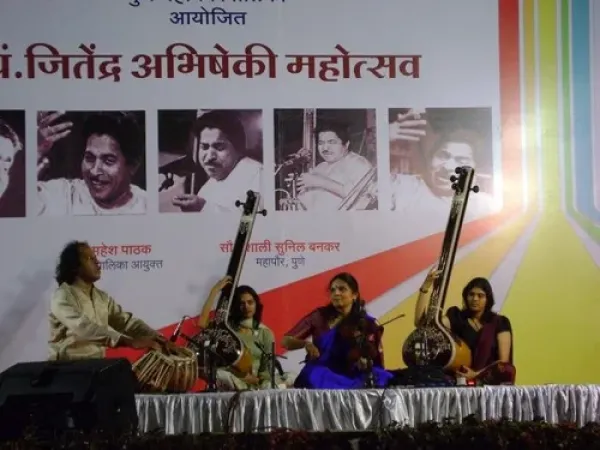 Four women performing classical Indian music on stage with traditional instruments, in front of a large banner with photos of prominent figures and text in Hindi.
