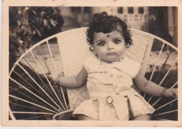 Black-and-white photograph of a young girl sitting on a woven outdoor chair with rounded back and armrests, outdoors with trees and a building in the background.