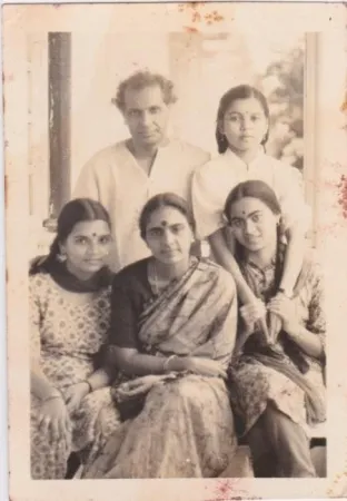 A black and white photo of five people, three women seated in front and a man and young girl standing behind them. They are indoors against a window and wallpaper backdrop, dressed in traditional Indian clothing.