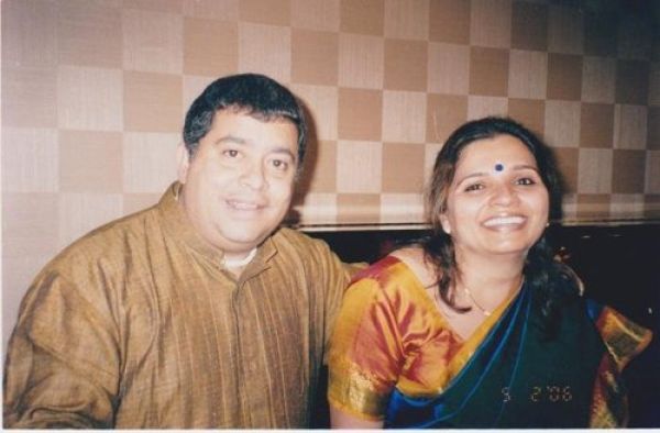 A man and woman sitting together, smiling for the camera in an indoor setting with a checkered wall background. The man is wearing a brown kurta, and the woman is dressed in a colorful saree with a bindi on her forehead.